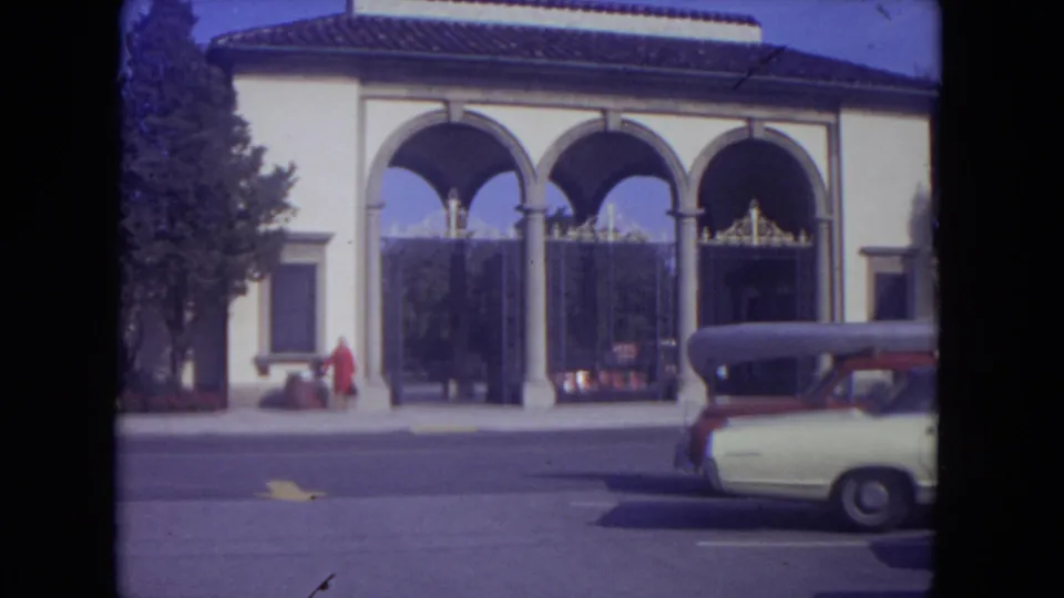 1972 8mm frame of the arched building entrance, with the woman in red moving or posing near the sidewalk and a car in front.