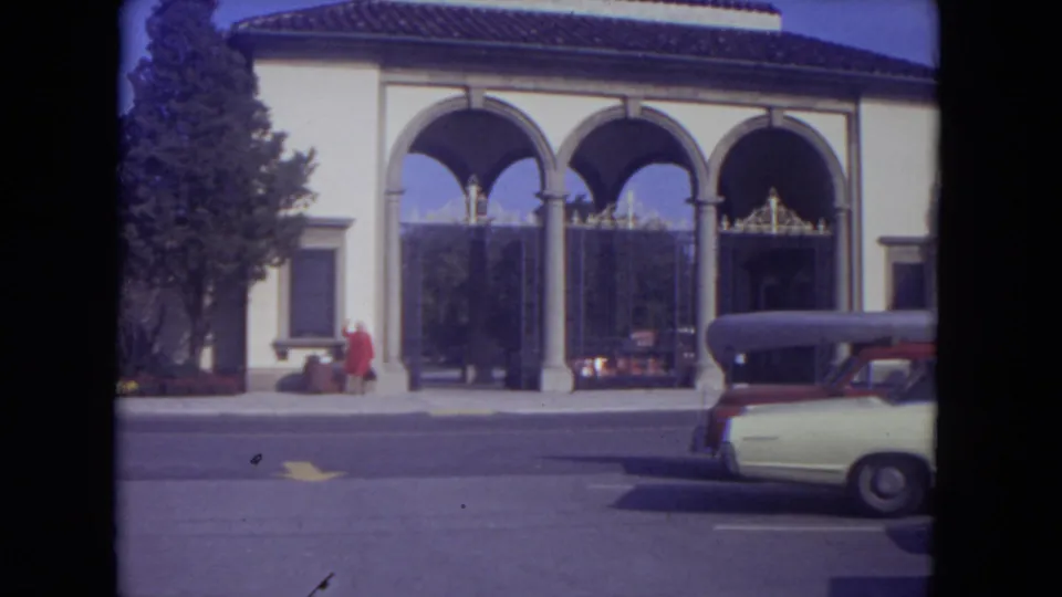 Lincoln Park 1972 home-movie view of a cream arched building with iron gates, a woman in red at the sidewalk, and a curbside car.