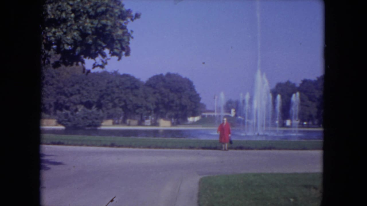 Woman in a red dress standing near a paved path with fountain spray behind her.