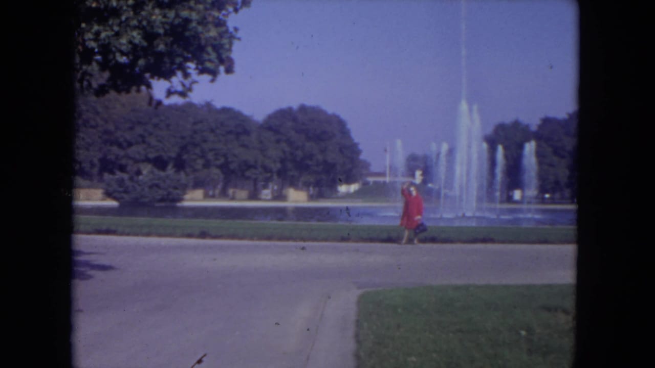 Woman in a red dress walking right along a path beside a large fountain.