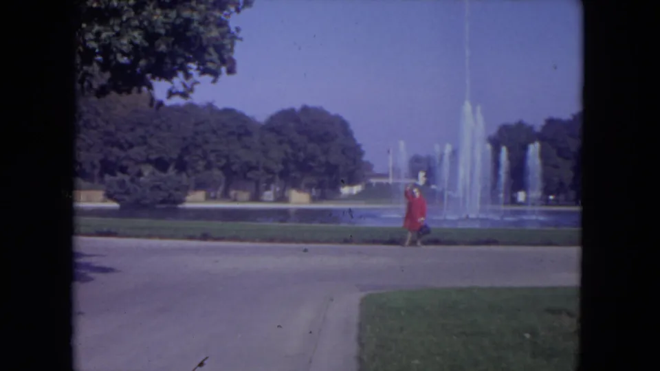 1972 Lincoln Park frame with the woman in red walking rightward along the paved path beside fountain jets and trees.