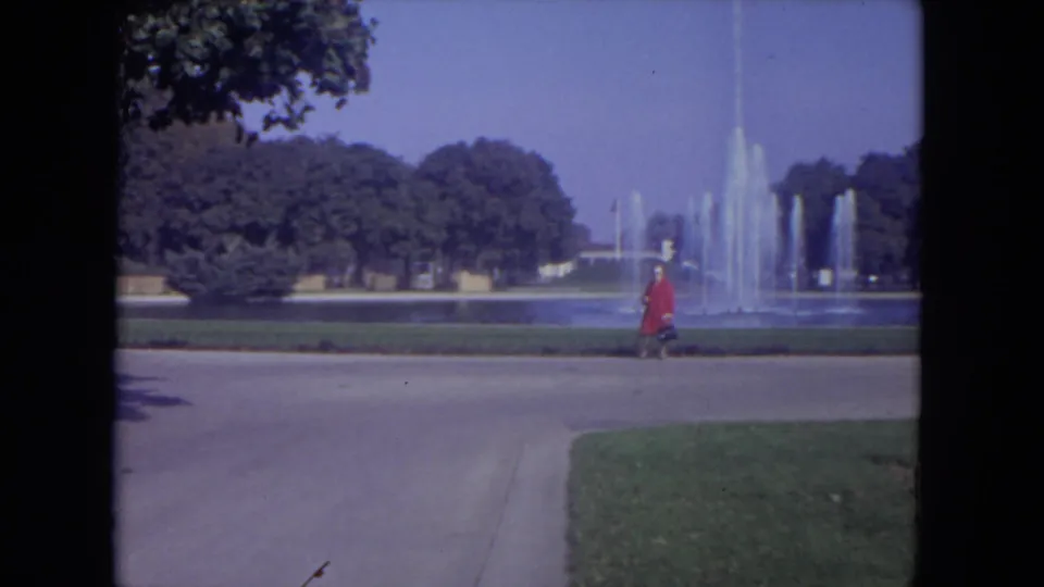 1972 Lincoln Park home-movie frame showing the red-dressed woman mid-walk before fountain spray, trees, and open lawn.