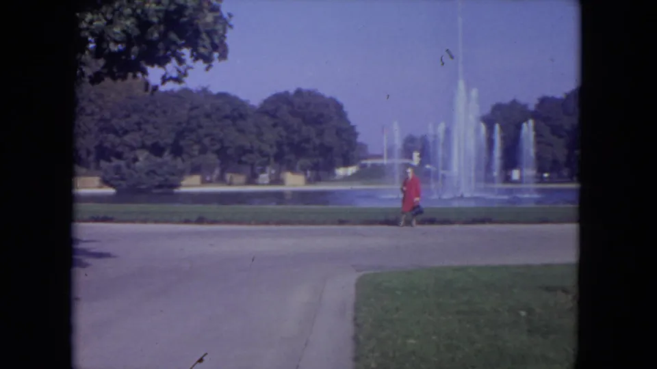 1972 Lincoln Park frame with the woman in red near the right edge, passing a fountain, lawn, and tree-lined background.