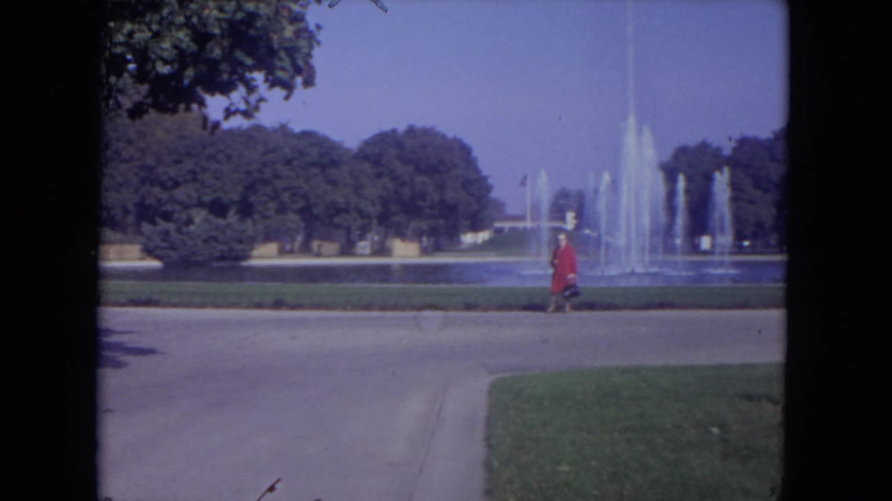 Woman in a red coat walks near a large fountain with trees and lawn behind her.