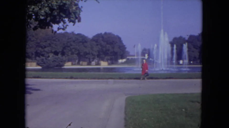 1972 Lincoln Park home-movie frame showing a woman in a red coat walking along a paved path beside an active fountain.