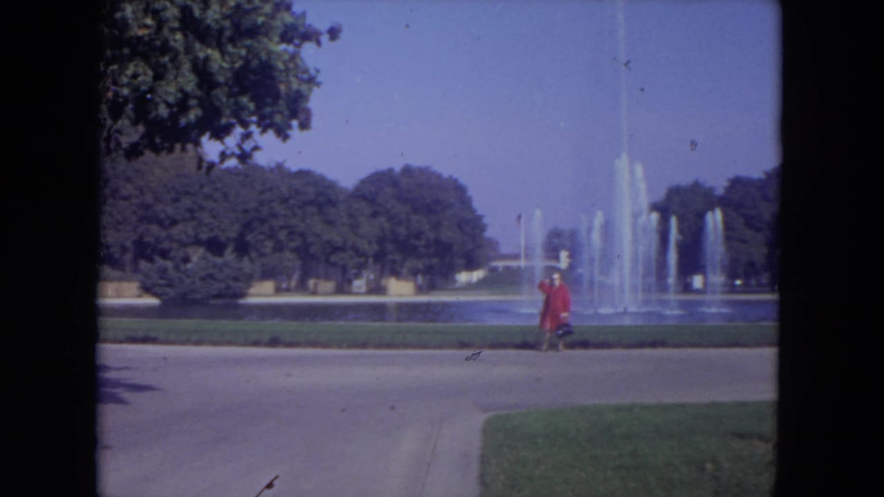 Woman in a red coat continues along the walkway with fountain jets in the background.