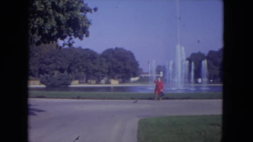 1972 Lincoln Park footage frame with a red-coated woman walking past fountain spray, lawns, and trees.