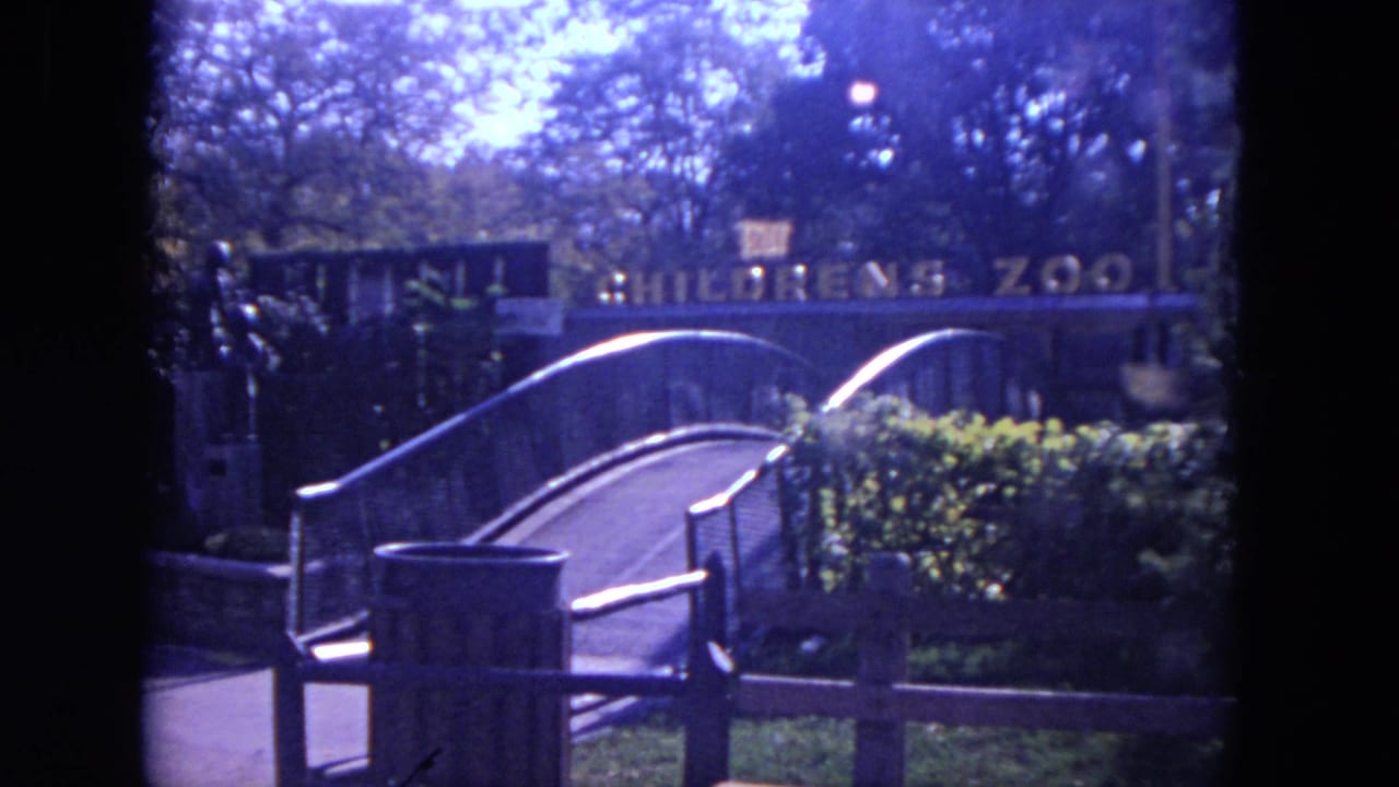 Curved bridge leading toward a Children's Zoo entrance sign behind railings and greenery.