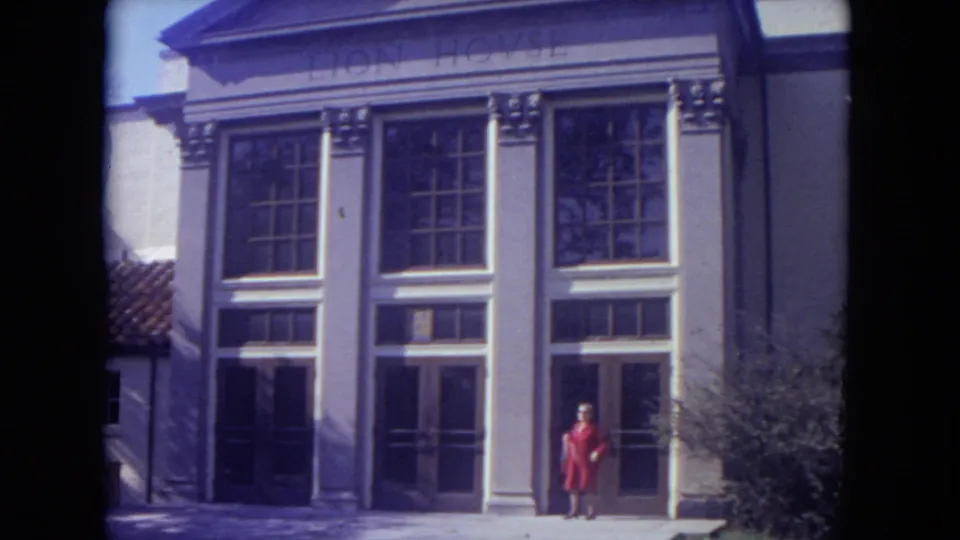 A woman in a red coat stands at the entrance of the Lion House facade in the supplied 1972 Lincoln Park zoo footage.