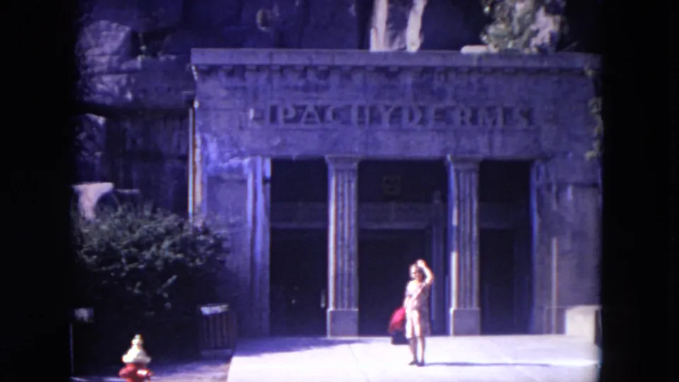 The woman waves from the walkway before the stone Pachyderms facade, with the entrance columns and shaded doorway behind her.