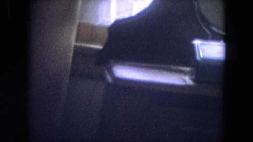 Dim 8mm interior frame, possibly inside a home or vehicle, with a dark human-like silhouette and seat or furniture shapes.