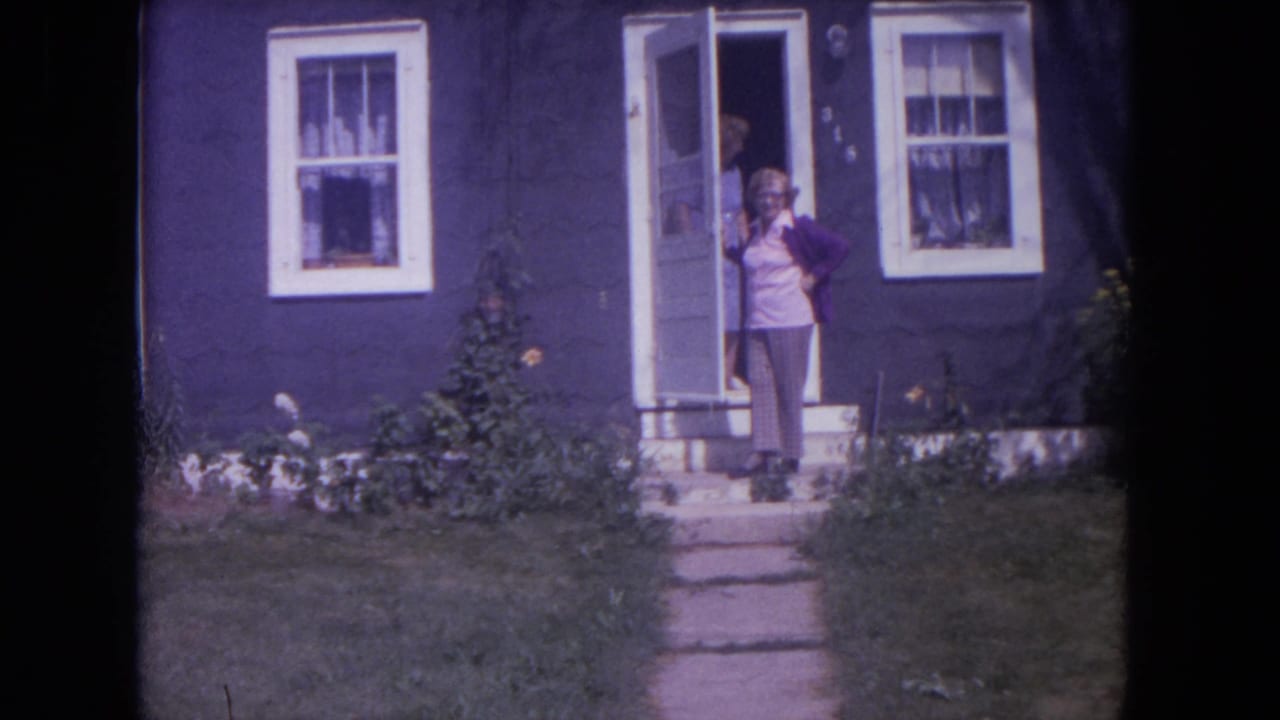 Woman stands on the front step of a dark blue Lake Geneva house, filmed in 1972 on 8mm.