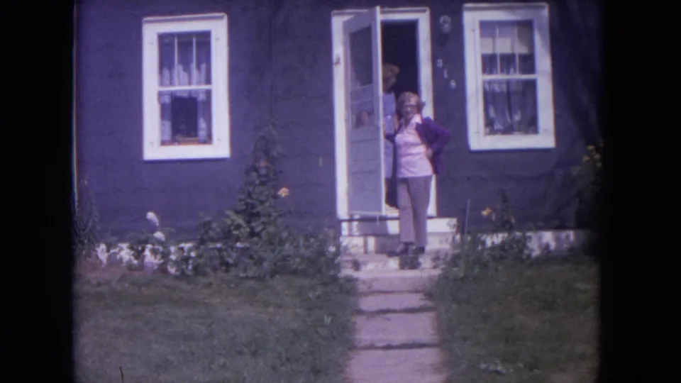 Woman remains near the open door of the house, with lawn and walkway visible in the 1972 Lake Geneva footage.
