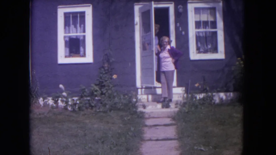 Woman stands at the open doorway of a dark blue house in Lake Geneva, Wisconsin, in 1972 home-movie footage.