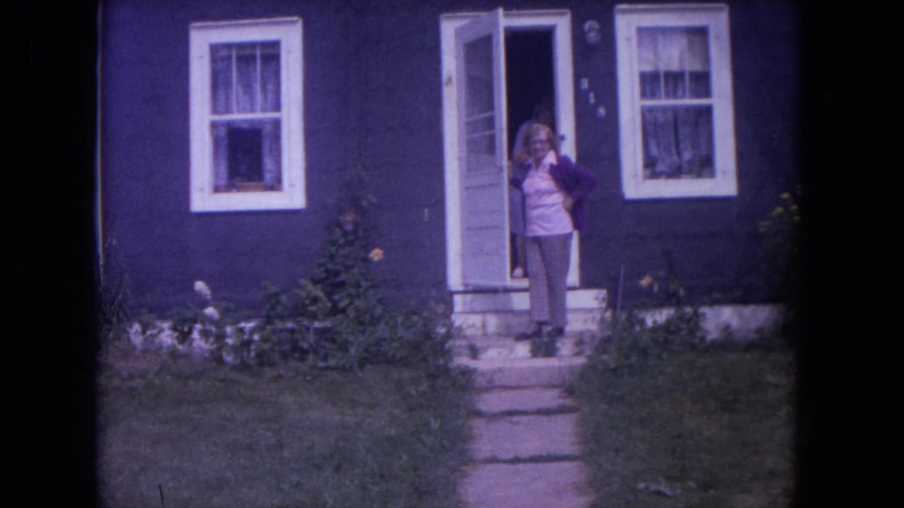 Woman in purple cardigan stands at the open front door of a dark blue house.