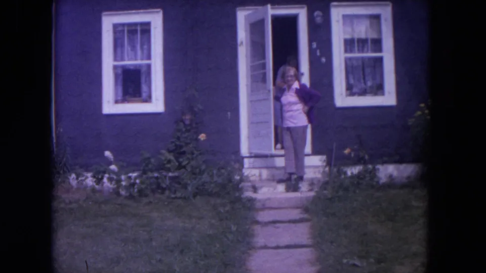 In 1972 Lake Geneva home-movie footage, a woman stands on the front steps of a dark blue house with white-trimmed windows.
