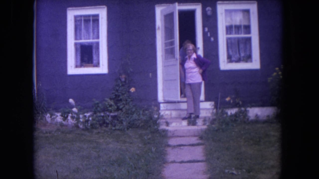 A woman stands near the doorway of a dark blue Lake Geneva house, framed by lawn, path, and white trim.