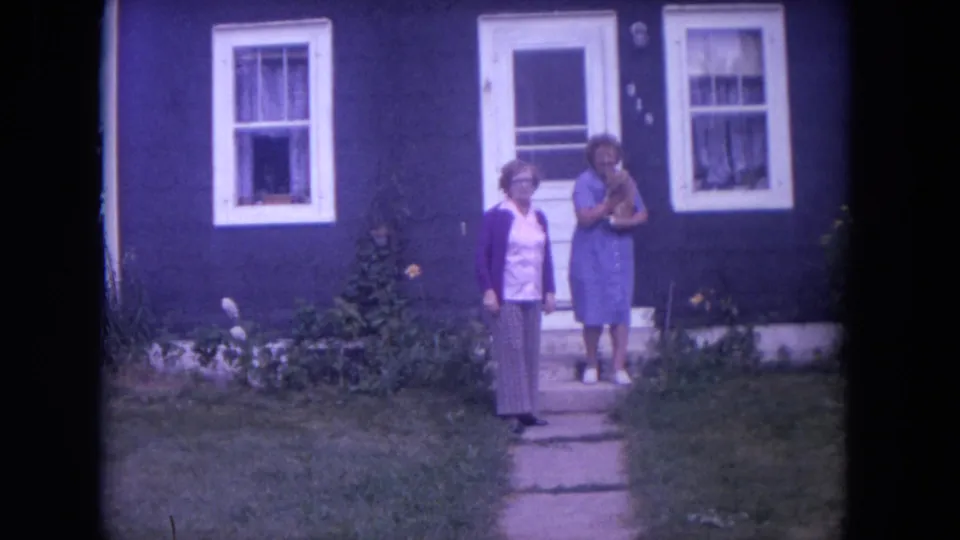 Two women stand outside the Lake Geneva house, one on the path and one near the doorway, in 1972 8mm footage.