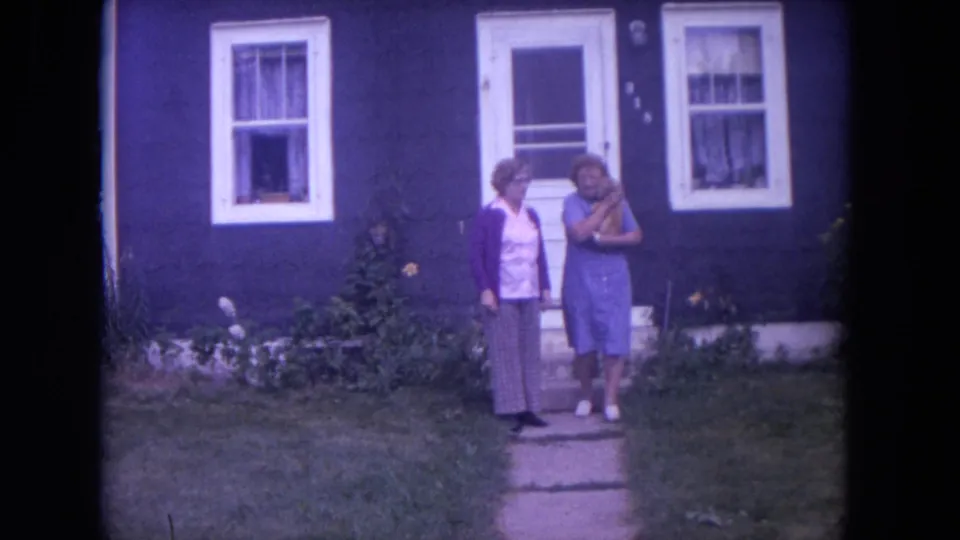 Two women stand together on the front path of a dark blue house in Lake Geneva, with white windows and garden plants behind them.