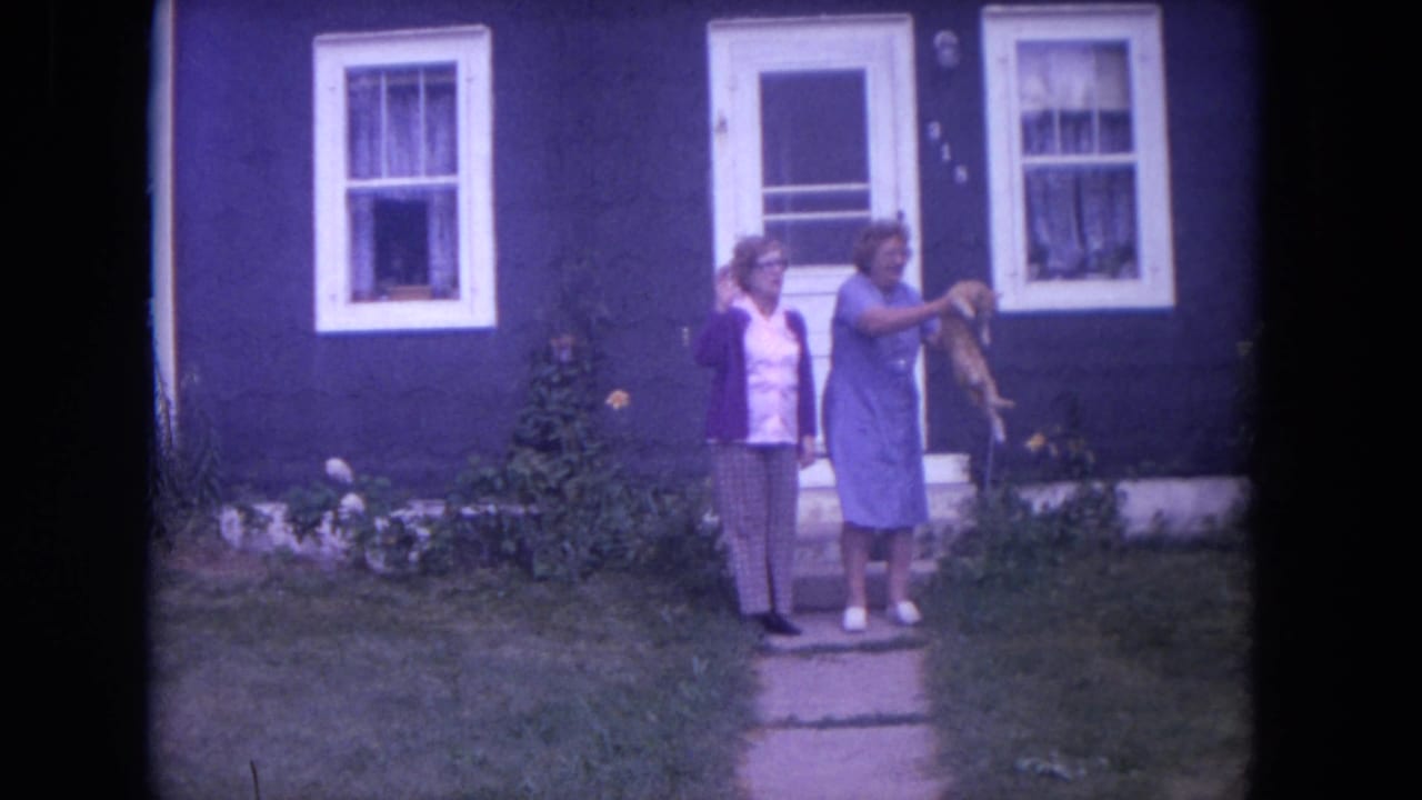 Two women stand on a sidewalk in front of a dark house, with one holding a cat.