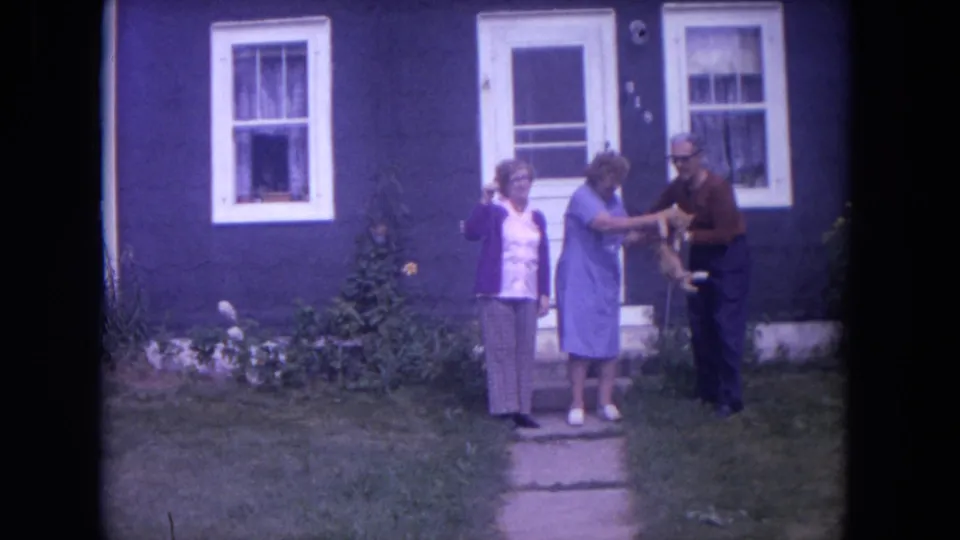 The man receives the cat from the woman in blue while another woman stands nearby in front of the house.