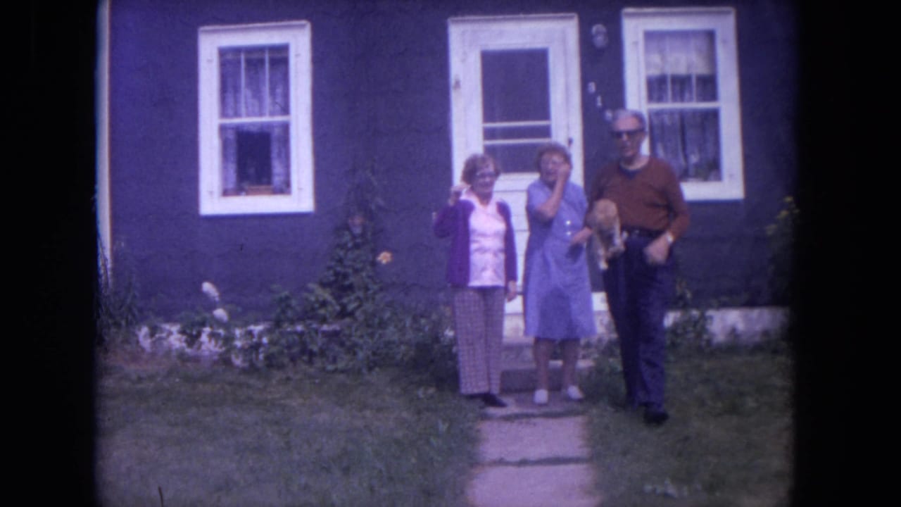 In 1972 Lake Geneva, Wisconsin, three adults pose outside a blue house with white trim as a man holds a ginger cat.