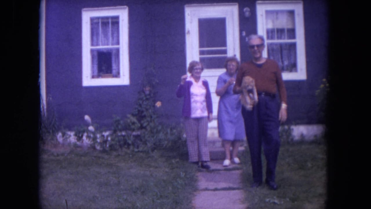 The man walks closer with the cat while two women remain near the house entrance.