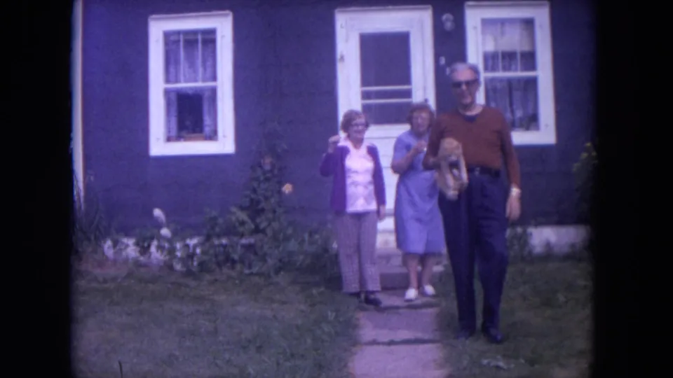 The man carrying the ginger cat steps toward the camera as two women stand by the blue house in Lake Geneva.