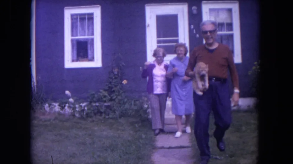 The family group begins moving toward the camera outside the blue house, with the cat visible in the man's arms.