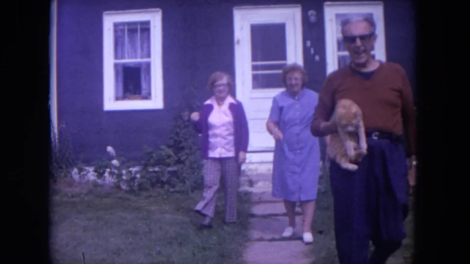 Near the end of the clip, the man with the ginger cat passes close to camera as two women follow outside the house.