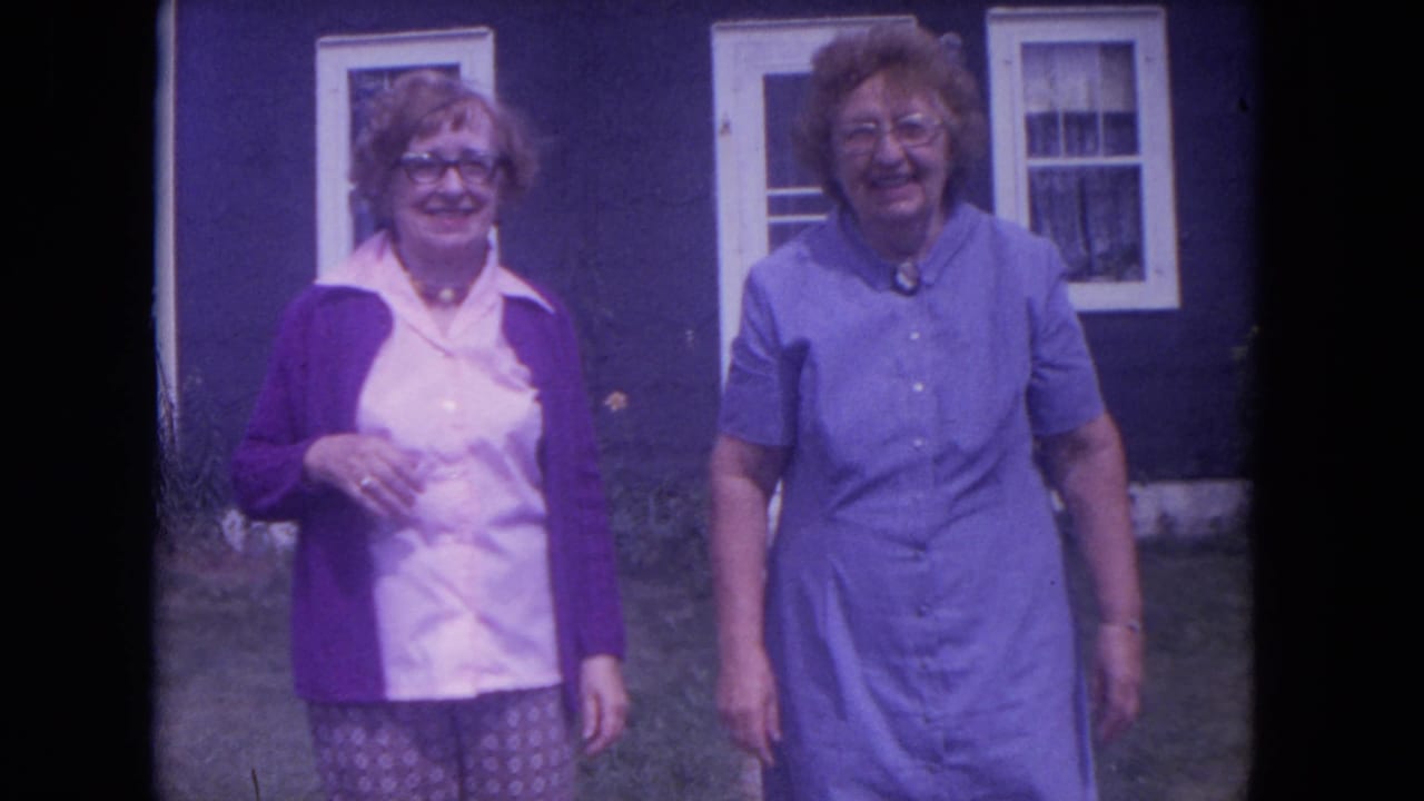 Two smiling women standing outside a purple house with white-trim windows.