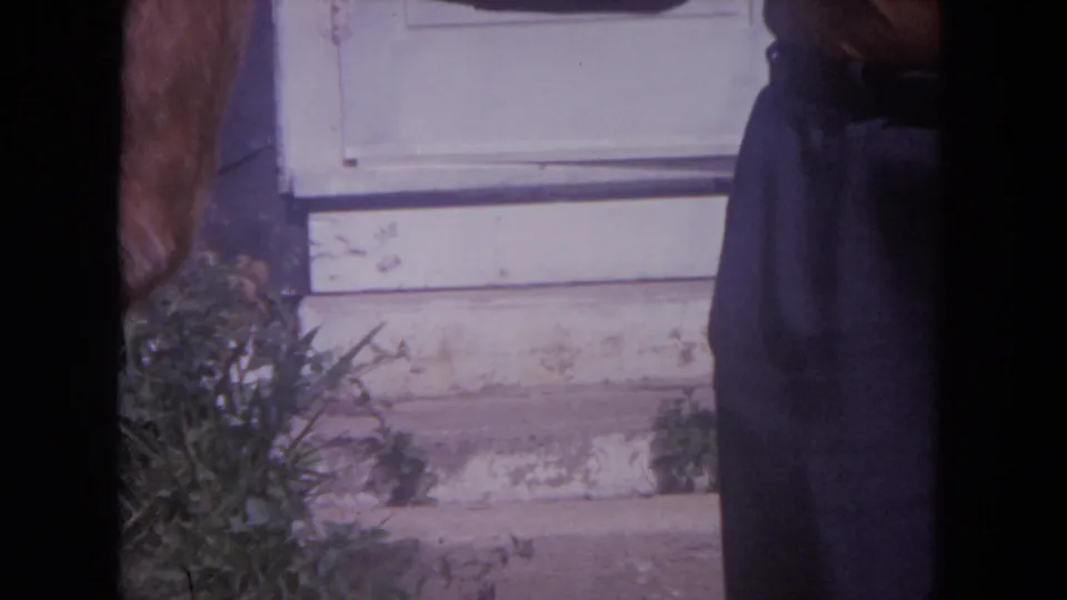 1972 Lake Geneva home-movie frame showing a white door, weathered step, foliage, and a partially cropped person at the right edge.