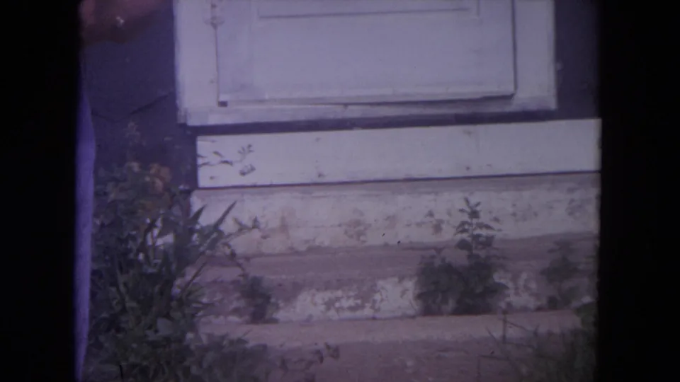 A 1972 Lake Geneva home-movie frame centers on a weathered white doorway, outdoor steps, plants, and a cropped person at the left edge.