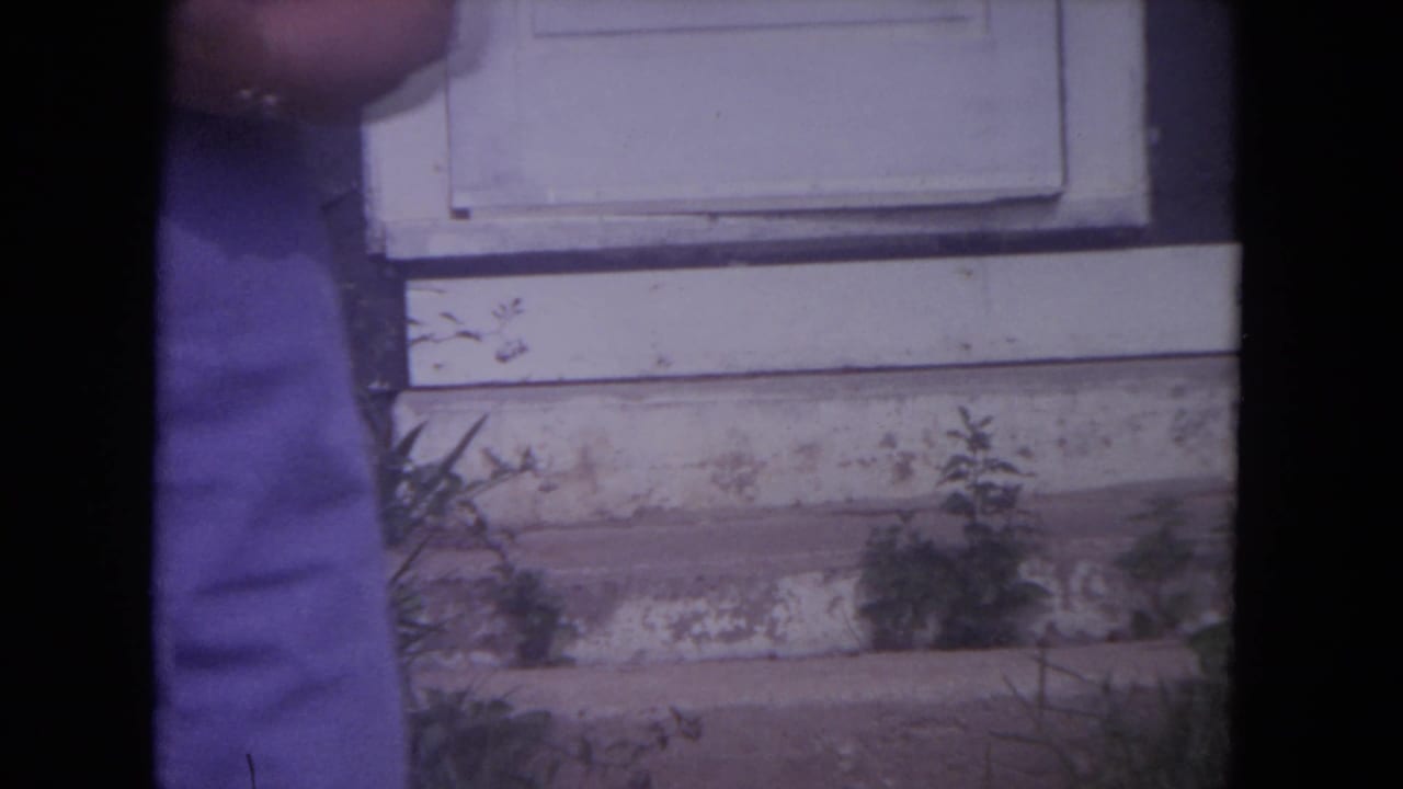 1972 Lake Geneva home-movie frame showing part of a woman standing near a weathered white door and exterior siding.