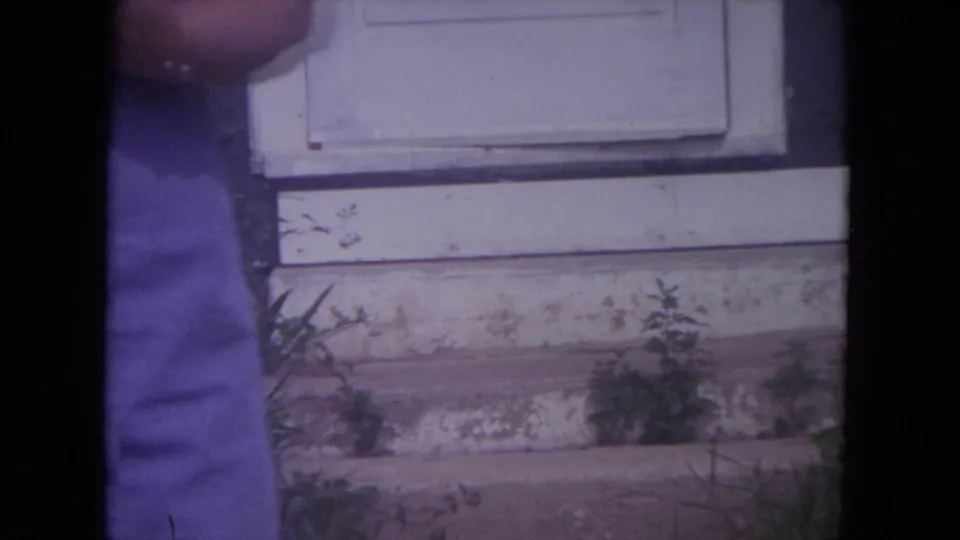 1972 Lake Geneva home-movie frame showing part of a woman standing near a weathered white door and exterior siding.