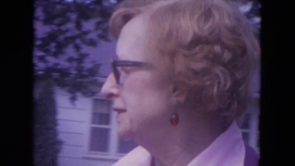 1972 Lake Geneva frame with the older woman in side profile, glasses visible, standing near white siding and a window.