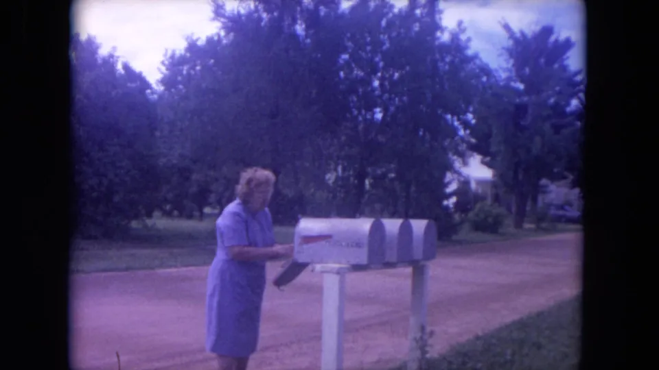 Older woman in a purple dress stands beside rural mailboxes on a dirt road in Lake Geneva, Wisconsin, in 1972 footage.