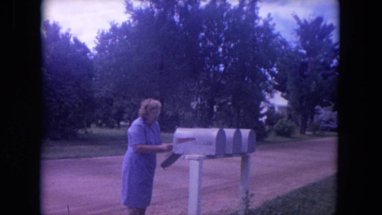 In 1972 Lake Geneva, Wisconsin footage, a woman in a purple dress appears to check a roadside mailbox near trees and a front lawn.