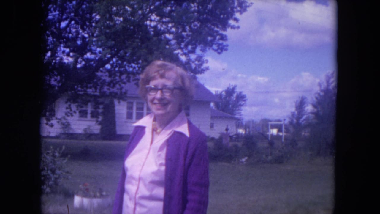Woman smiling in a front yard with a white house and trees behind her.