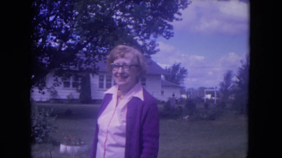 Closer 1972 home-movie frame of the woman smiling in a Lake Geneva yard, with a white house, lawn, and large tree visible behind her.