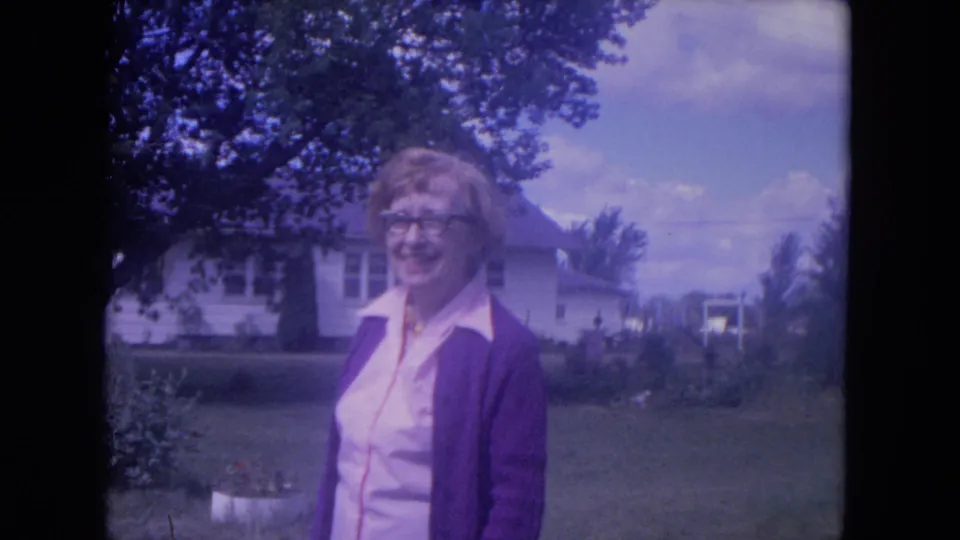 The woman remains smiling in the Lake Geneva residential yard, with 8mm color shift, a white house, trees, and garden details behind her.