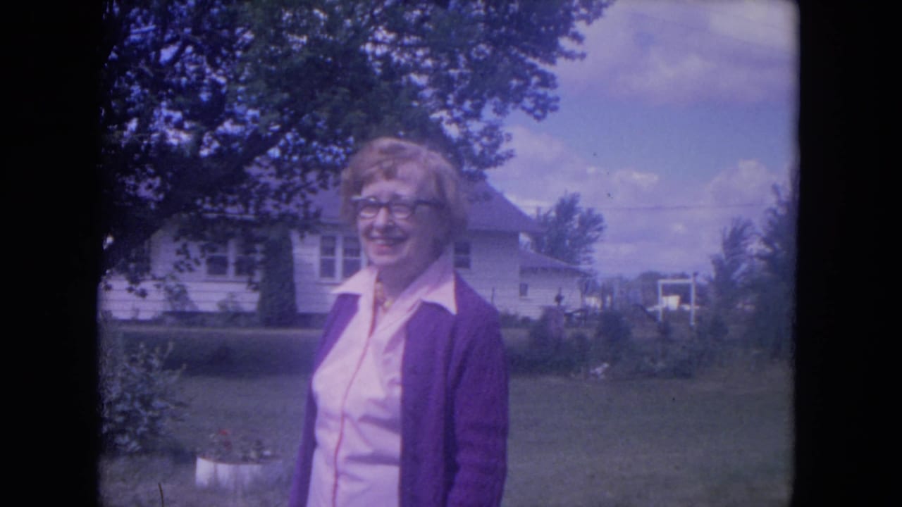 In 1972 Lake Geneva footage, a woman in glasses and a purple cardigan smiles outdoors near a lawn, trees, and a white house.