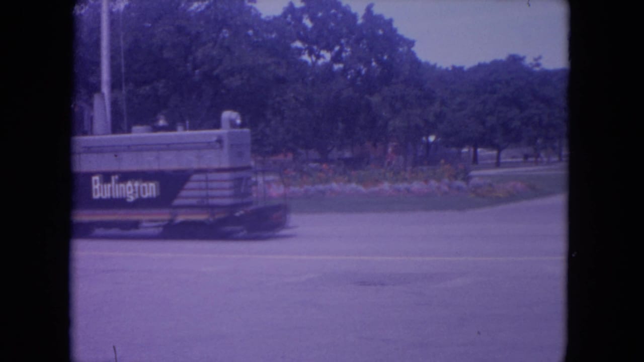 Small train marked Burlington moves along a paved street beside trees and flower beds.