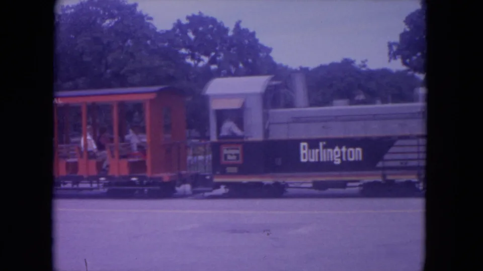 A red passenger car with visible riders follows the Burlington-marked train in 1972 Lake Geneva park footage.
