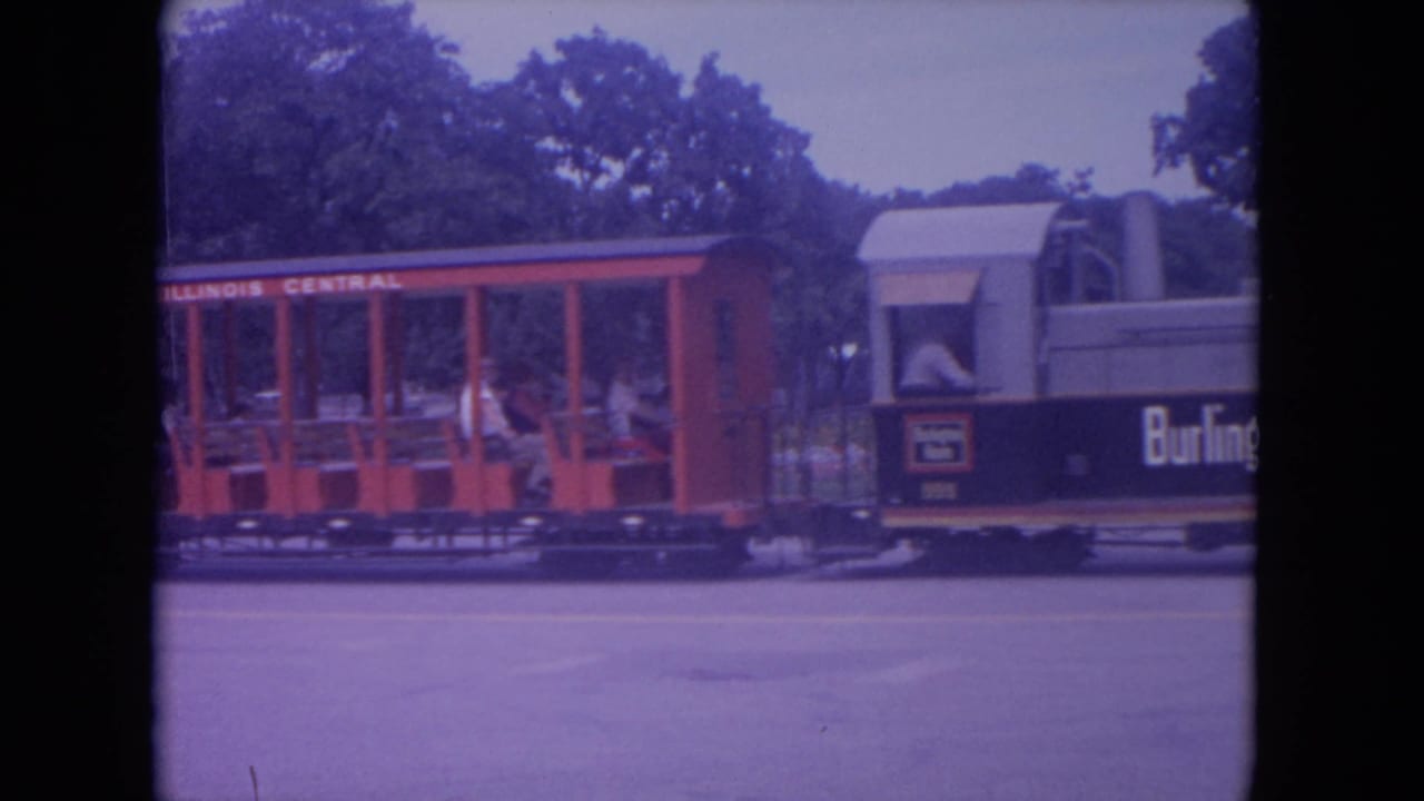 Red open-air Illinois Central park train car with riders passing trees.