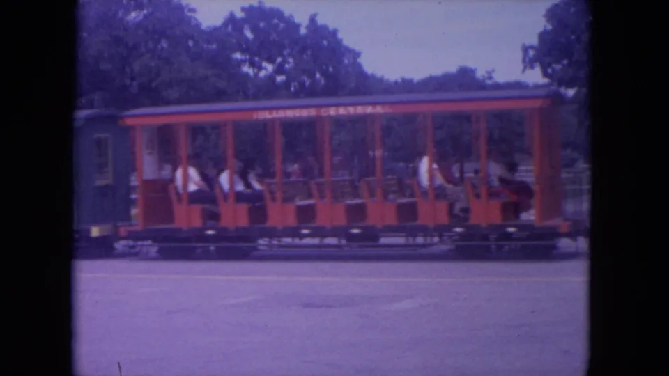 1972 Colby, Wisconsin frame of a red Illinois Central open passenger car carrying riders along a park track.