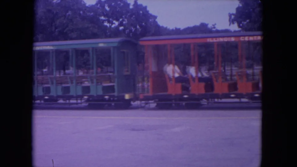 1972 Colby, Wisconsin 8mm frame showing green and red open-air park train cars with passengers seated inside.