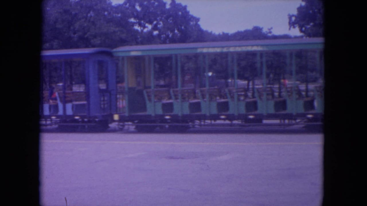 Open-sided green and blue children's train car passing on pavement with riders visible inside.