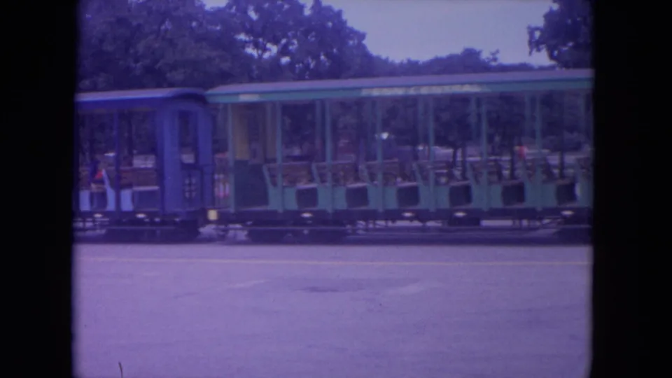 1972 Colby, Wisconsin home-movie frame showing a green and blue open-sided children's train car moving past trees and pavement.