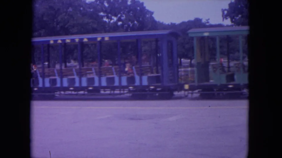 The open children's train continues past the camera in Colby, Wisconsin, with riders seated under a green roof in a 1972 8mm frame.